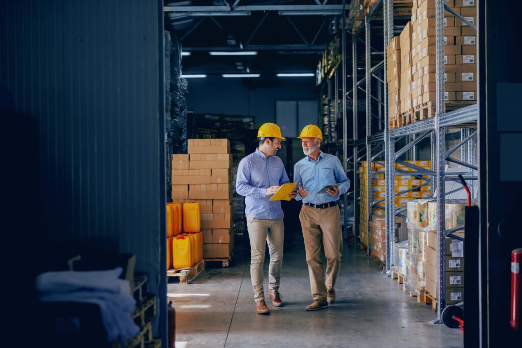 two men wearing khaki pants and blue button down shirts walk through a manufacturing facility wearing yellow hard hats and having a conversation